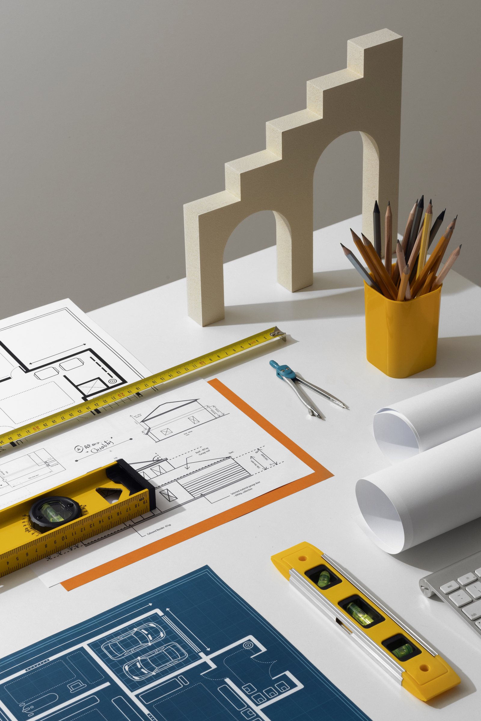 Overhead view of an architectural desk with a stepped-arch scale model, pencils, rolled blueprints and floor plans