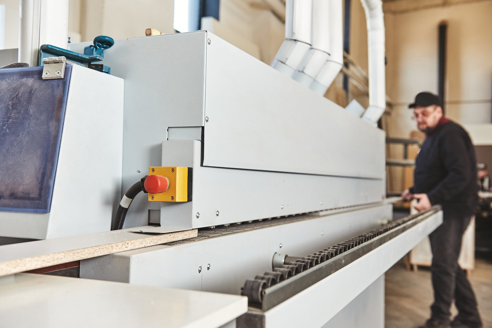 Edge-banding machine processing a panel, worker adjusting settings in the background