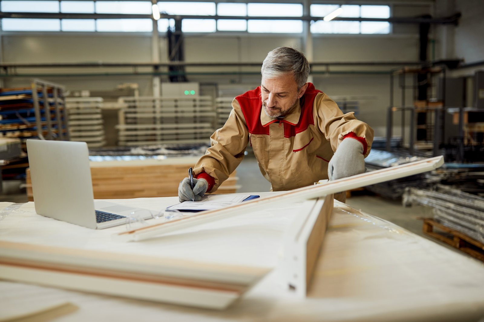 Worker noting measurements against a clipboard beside cut boards and a laptop in a workshop