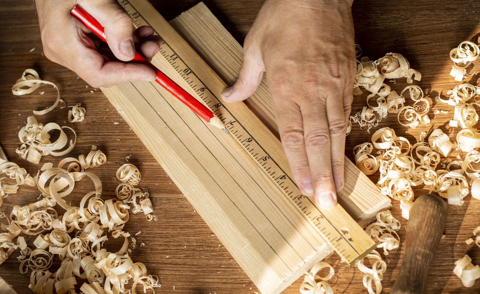 Hands marking a wood plank with a ruler and pencil, curled wood shavings scattered around