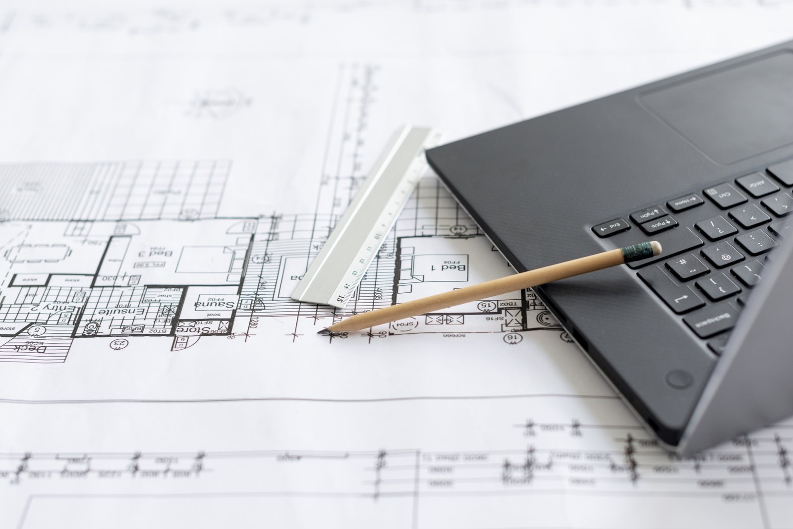 Floor plan blueprint with a metal ruler, pencil and the corner of a laptop keyboard laid across it