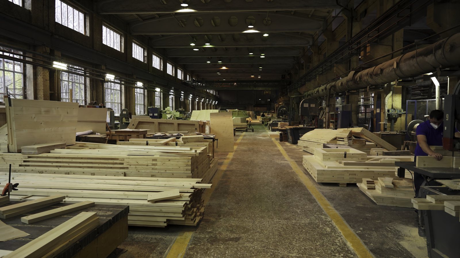 Woodworking shop floor with stacked lumber and workstations along both sides