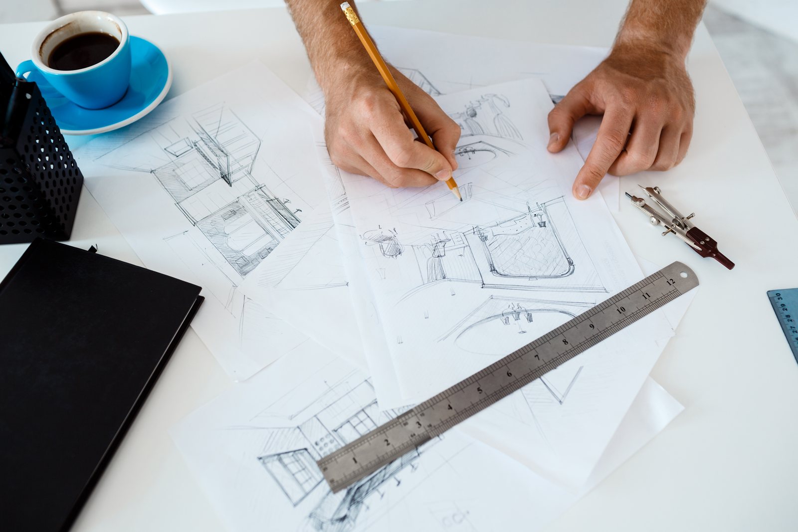 Hands sketching a floor plan with a ruler and compass at a desk, coffee alongside