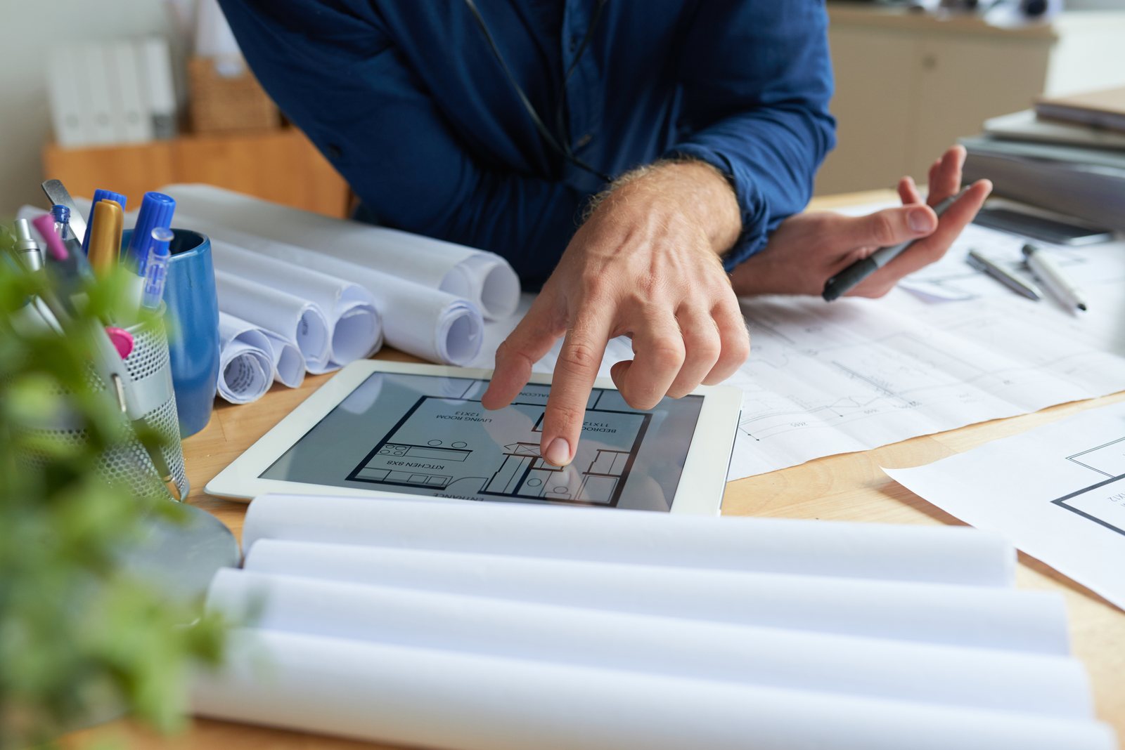 Person reviewing a floor plan on a tablet at a desk with rolled drawings and pens